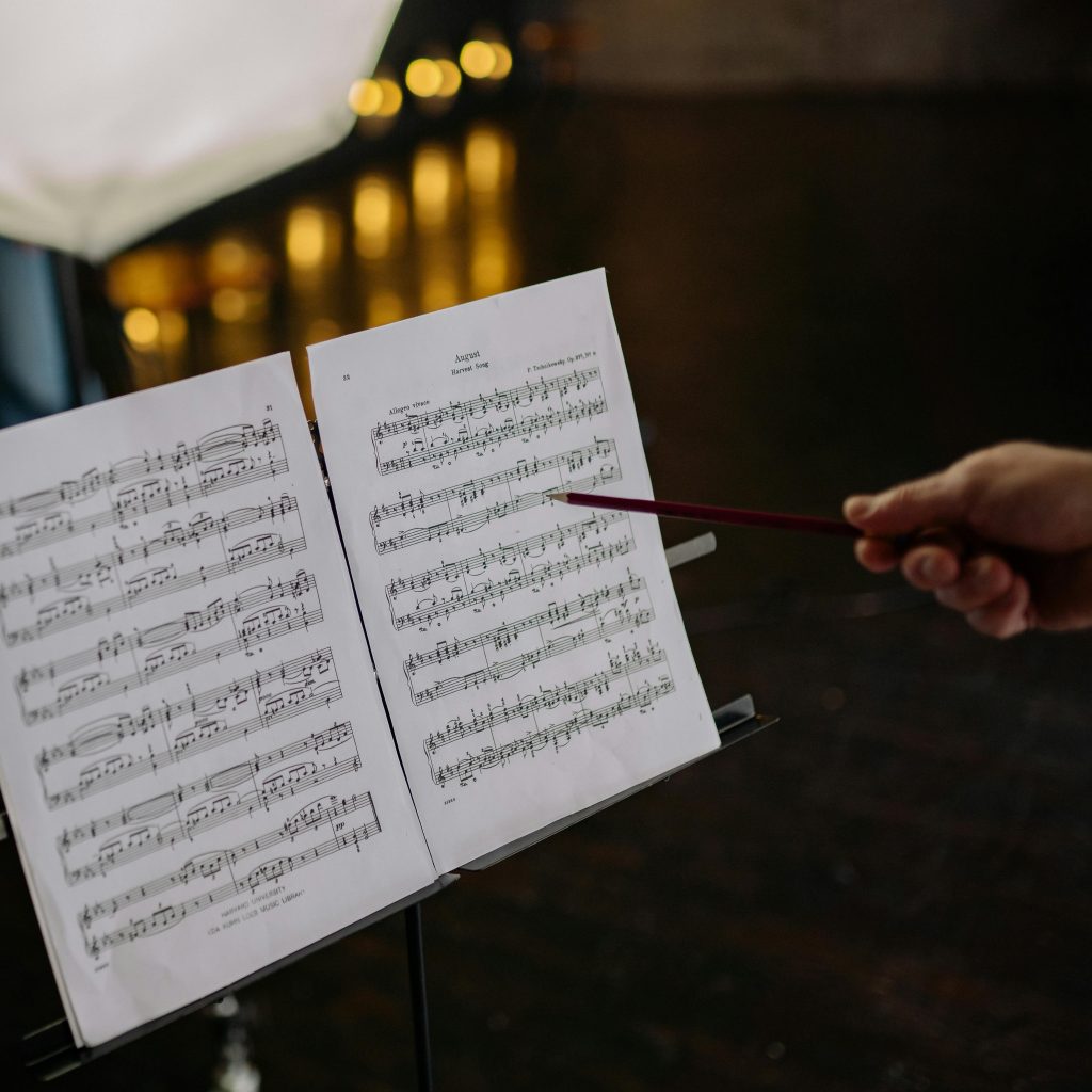 Close-up of an instructor pointing at sheet music on a stand, indoors, with blurred lights in the background.
