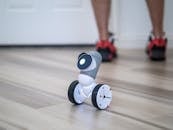 A close-up of a modern wheeled robot on a wooden floor with legs in the background.